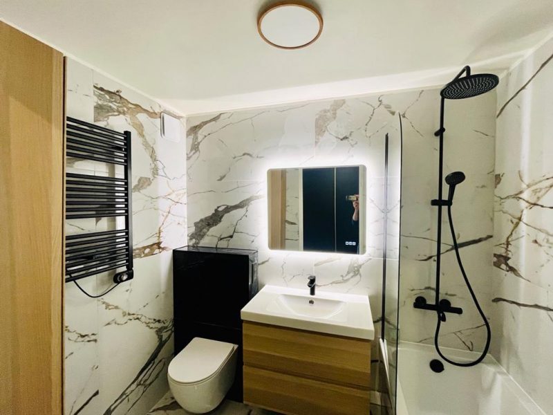 photo du projet de rénovation, montrant une salle de bains avec une majorité de marbre blanc associé à un coffrage en marbre noir , un meuble vasque et une colonne en bois et des équipements sanitaire en metal noir